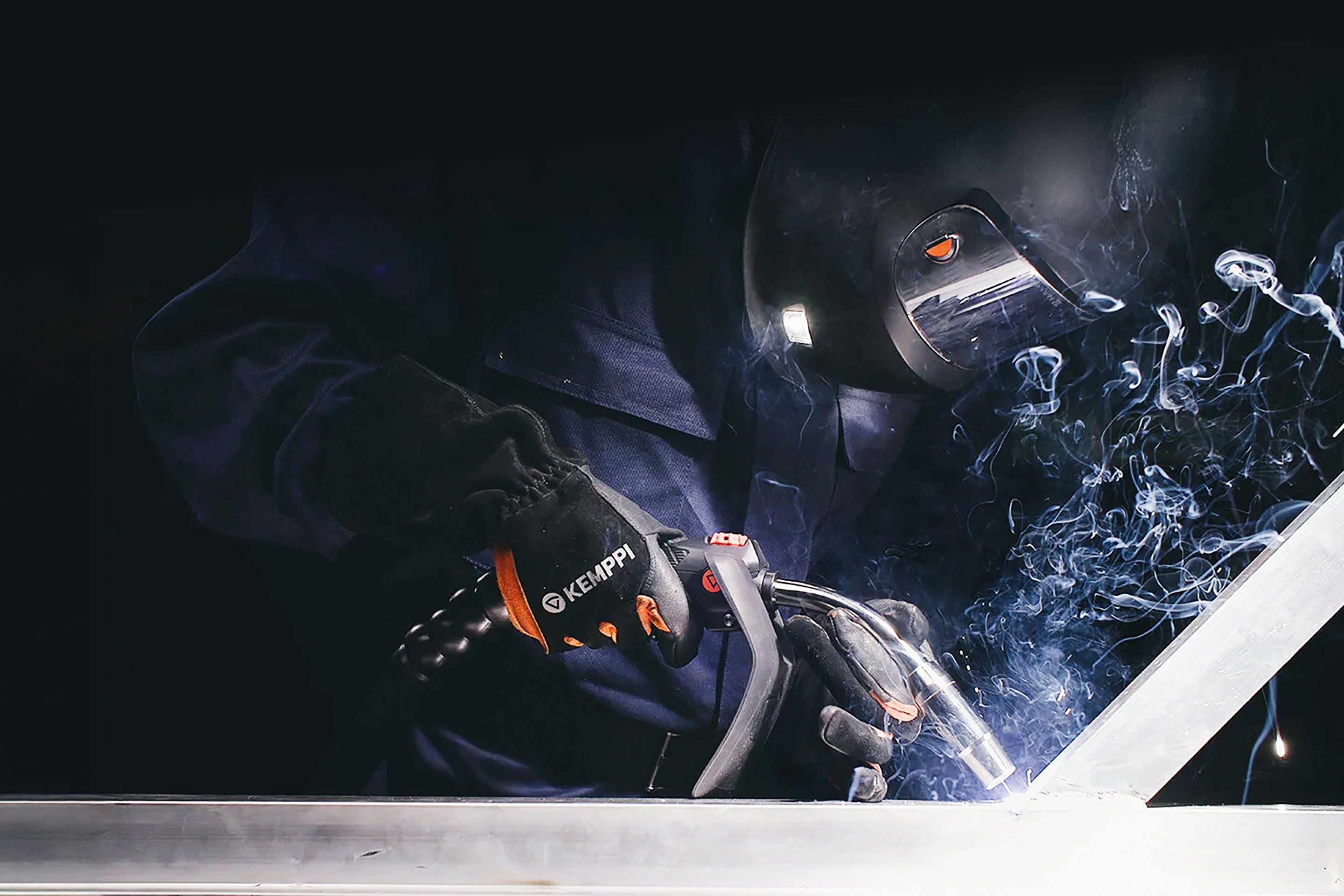 A close-up of a person welding metal in a dark environment with smoke rising from the weld, wearing protective gloves and a helmet, with the word 'KEMPPI' visible on their gear.