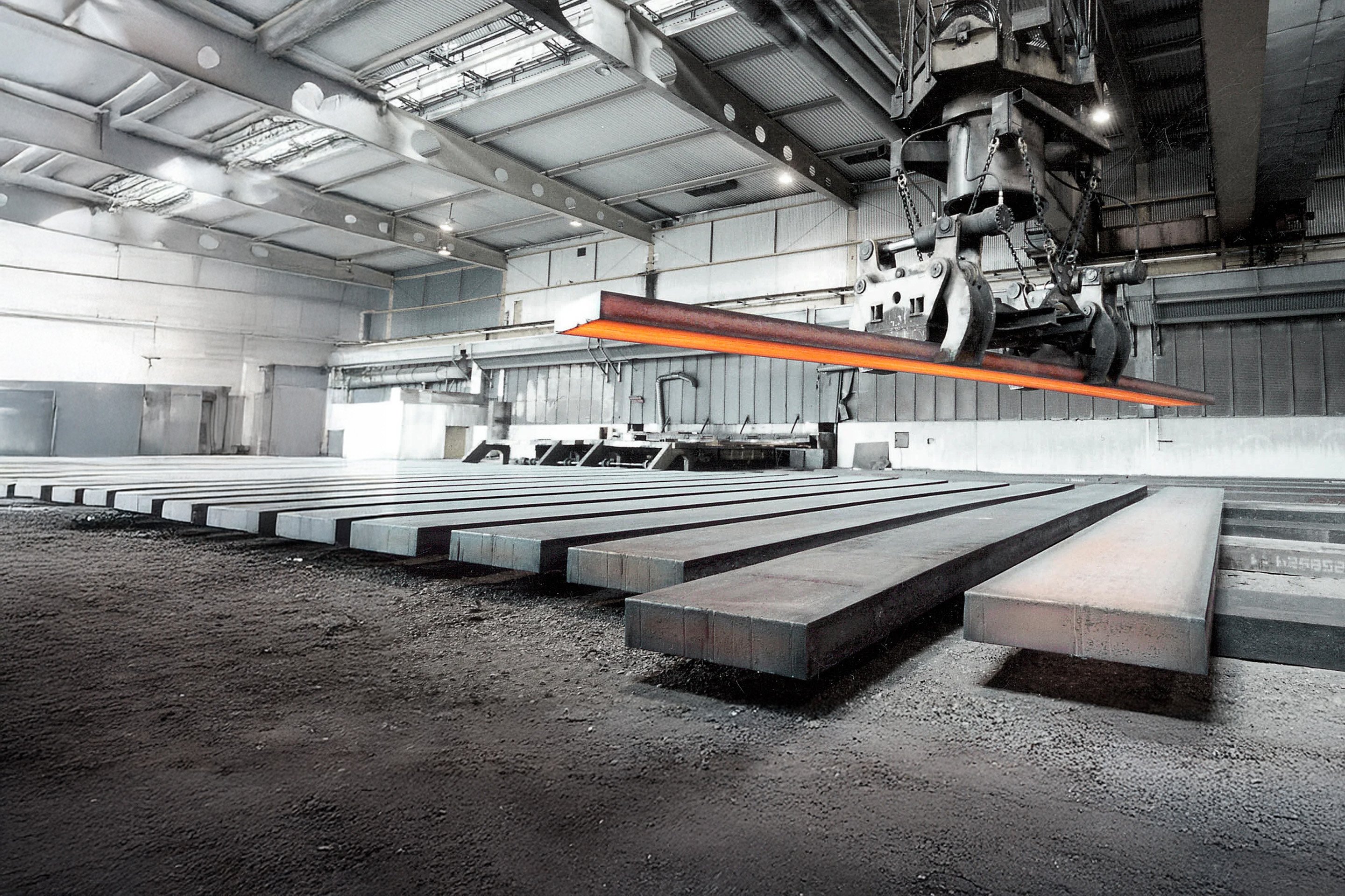 Industrial steel manufacturing facility with large steel slabs on the ground and a crane lifting a glowing piece of steel in a large, dimly lit warehouse.