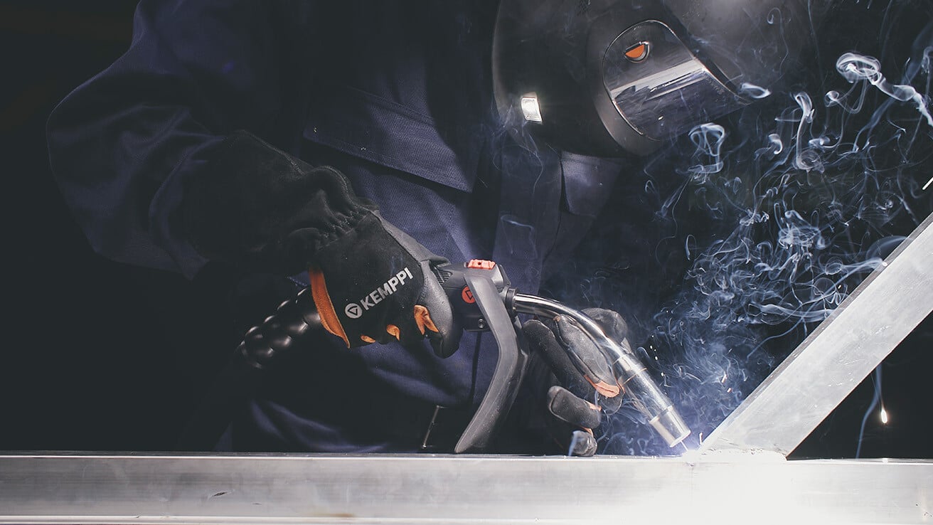A welder wearing a helmet and gloves with the 'Kemppi' logo, using a welding torch to work on metal, surrounded by smoke from the welding process.