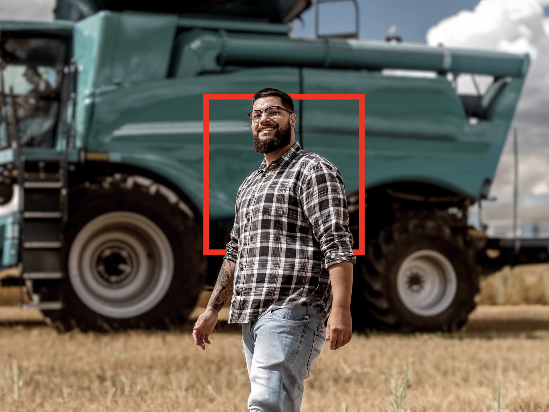 Man with glasses and beard standing confidently in front of a large green farming vehicle, wearing a plaid shirt and jeans.